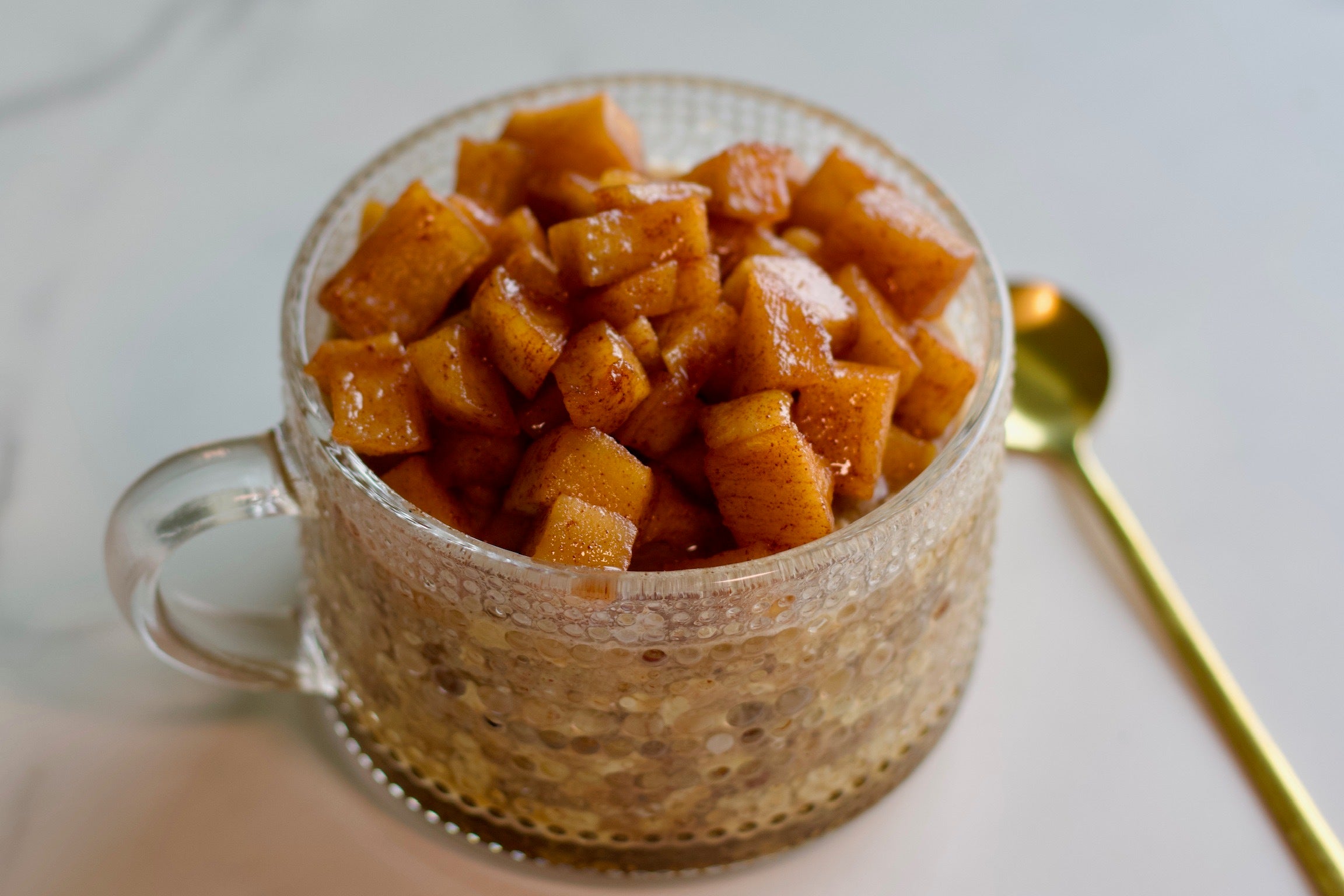 A glass mug of creamy JOI Apple Pie Overnight Oats topped with caramelized apple chunks and a gold spoon.