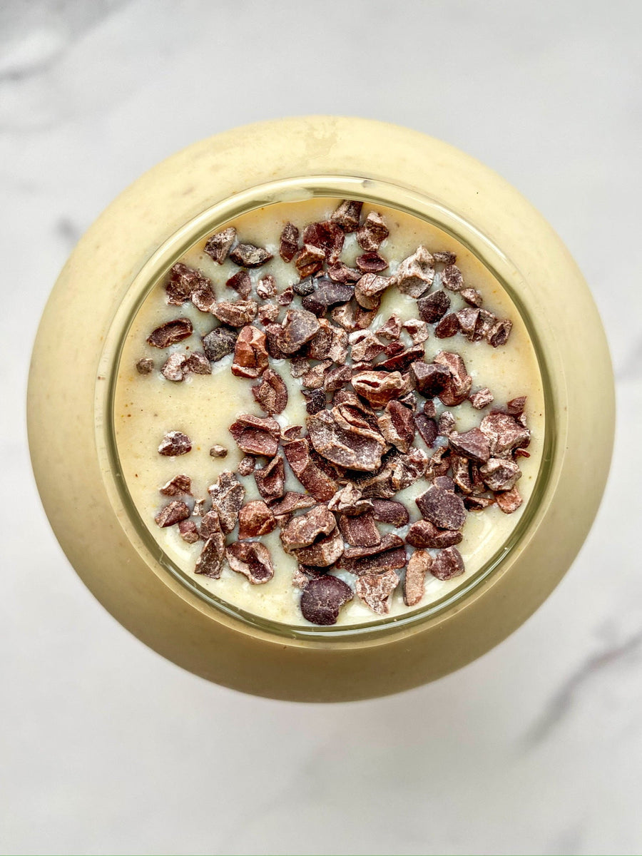 An overhead view of a banana smoothie with cacao nibs, made with JOI plant base, in a glass against a marble background.