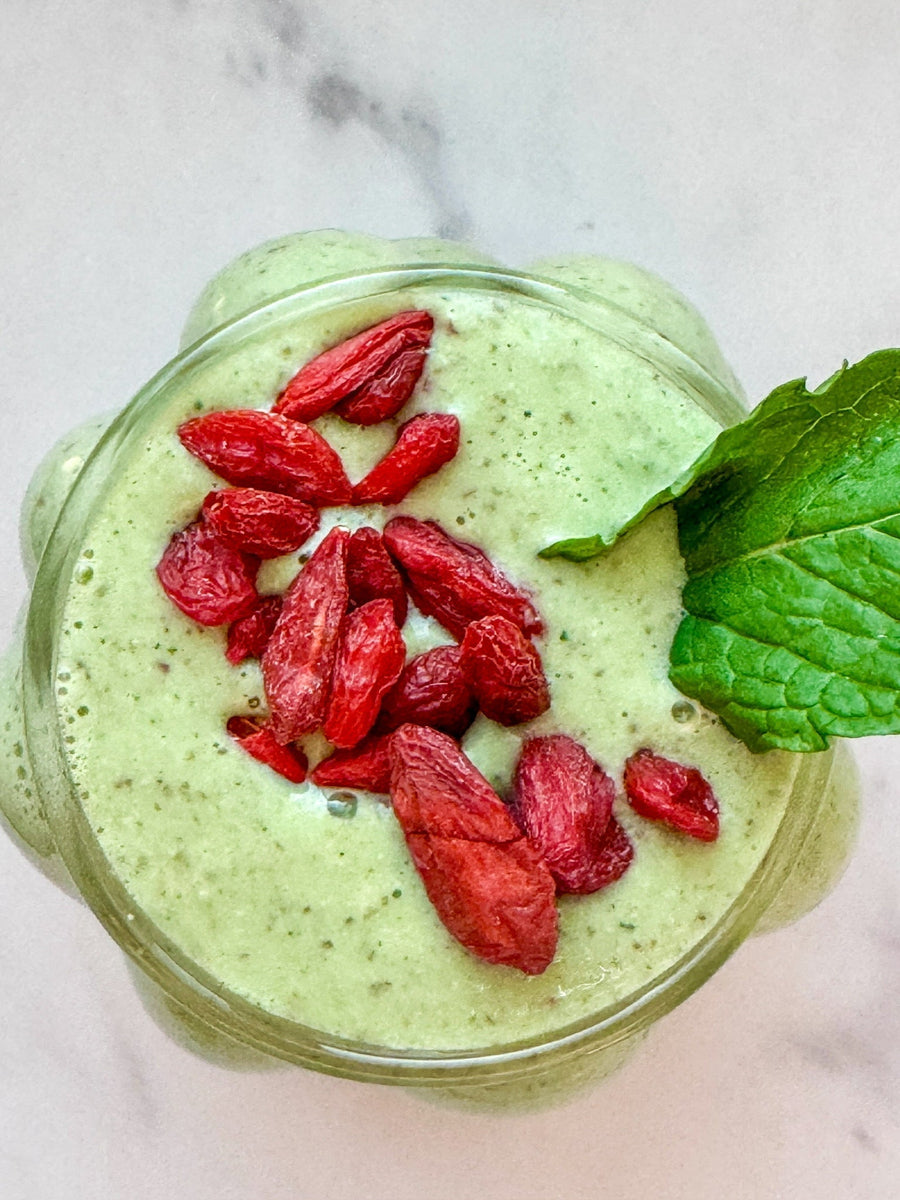 A vibrant green JOI smoothie, topped with dried goji berries and a fresh mint leaf, on a white marble surface.