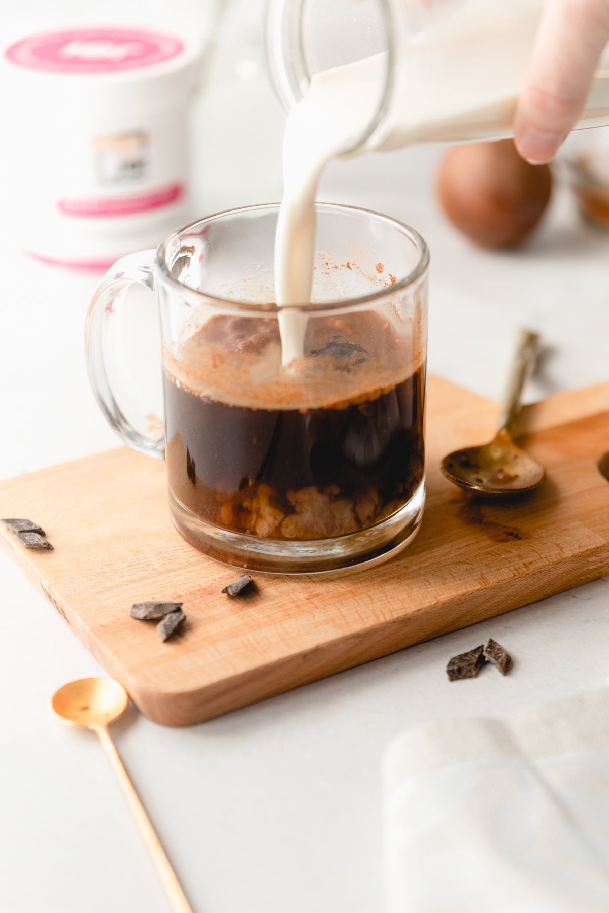 Plant-based JOI creamer is poured into a glass mug of dark coffee, sitting on a wooden board with chocolate pieces.