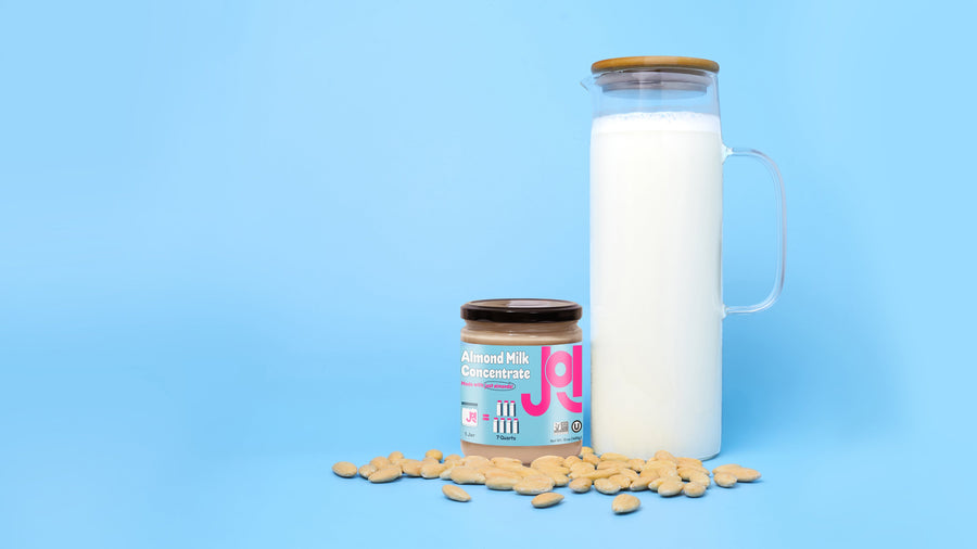 A glass pitcher of JOI plant-based almond milk stands next to a jar of almond milk concentrate and loose almonds.