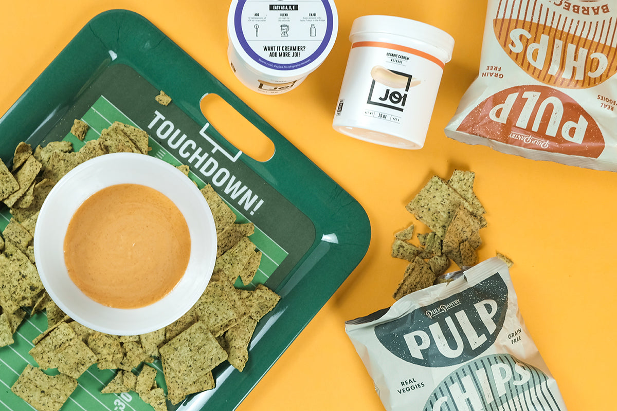 JOI plant bases and Pulp Chips bags are surrounded by green square chips and a bowl of vegan buffalo hot sauce dip.