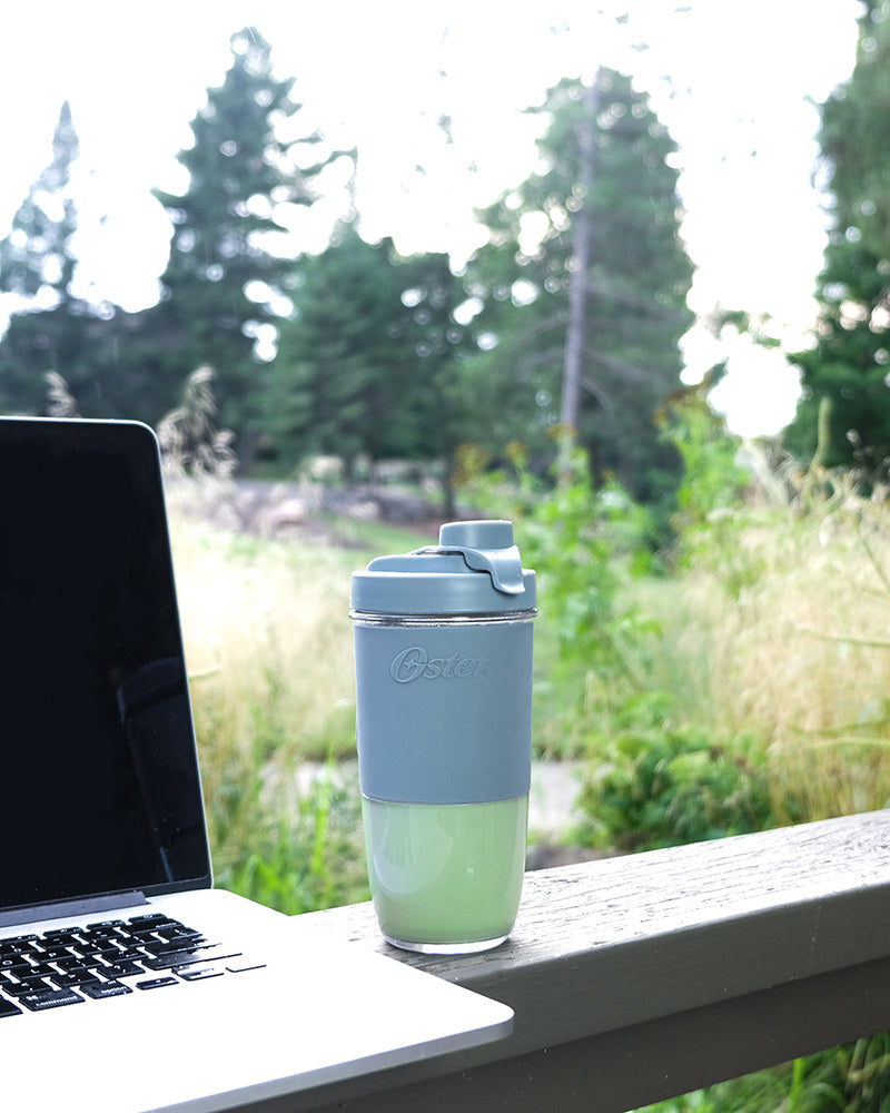 An Oster travel mug containing a JOI Spiced Vanilla Matcha Latte sits next to a laptop outdoors.