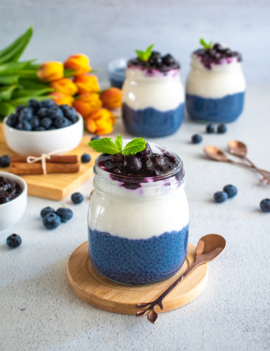 A jar of JOI chia pudding with a blue layer topped with white yogurt and blueberry compote, alongside fresh blueberries and orange tulips.