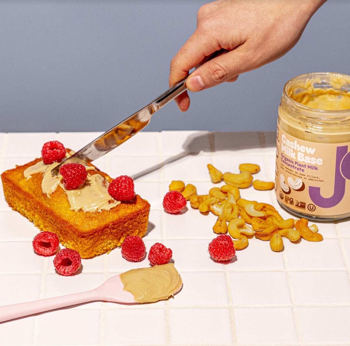 A hand spreading JOI Cashew Milk Base on a loaf cake with raspberries, surrounded by cashews and a jar.