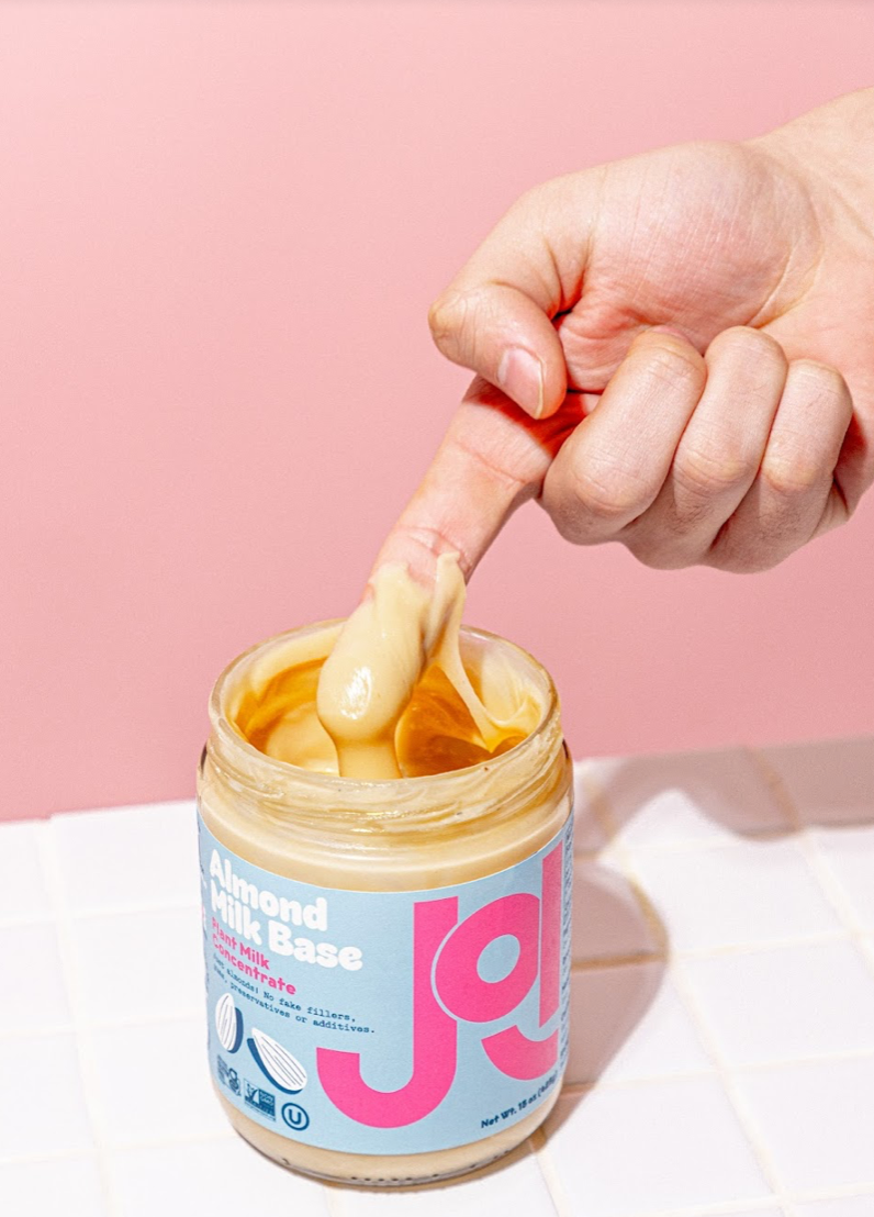 A finger dips into a jar of JOI Almond Milk Base concentrate, which is displayed on a white tiled surface.