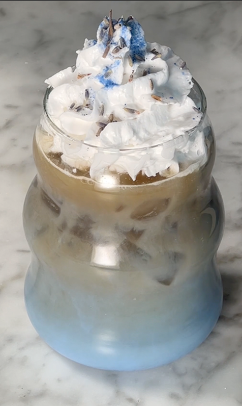 An iced lavender oat milk latte, made with JOI plant base, topped with whipped cream, lavender buds, and blue sprinkles.