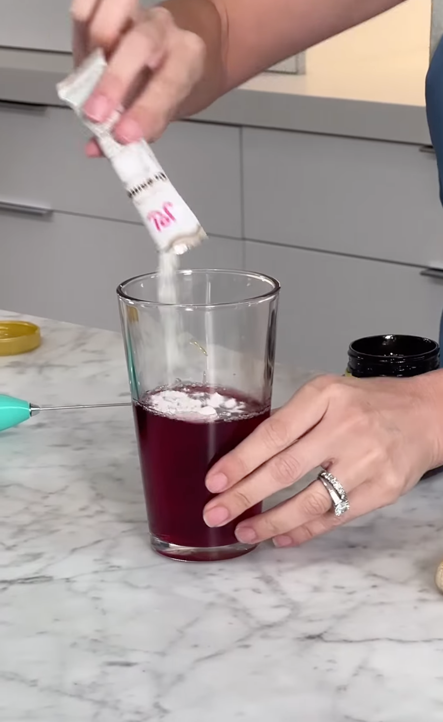 A hand pours white JOI oat milk powder from a packet into a glass of magenta liquid, next to a teal frother.