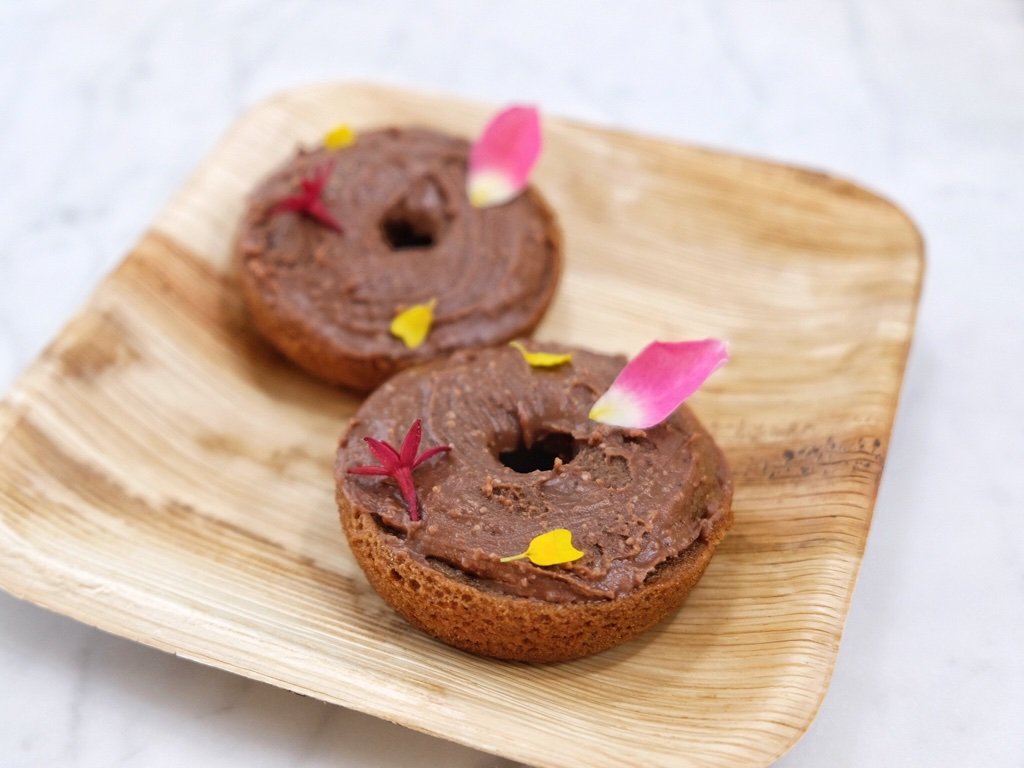 Two JOI-based chocolate donuts, topped with chocolate frosting and garnished with small colorful petals, on a wooden plate.