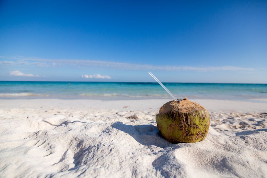 A green coconut with a straw sits on white sand at a beach, representing the benefits of coconut water.