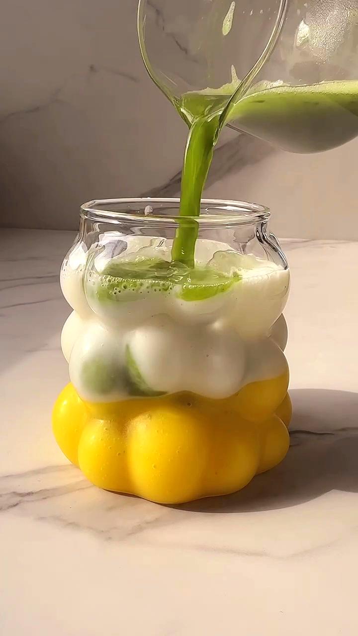 Green matcha JOI plant milk is poured into a cloud-shaped glass with white foam and yellow mango.