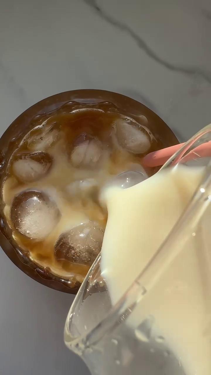 Rich JOI almond coffee creamer is poured from a glass pitcher into an iced coffee with a pink straw.