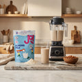 A JOI Almond Milk Powder bag sits next to a blender and glass of prepared almond milk in a kitchen.