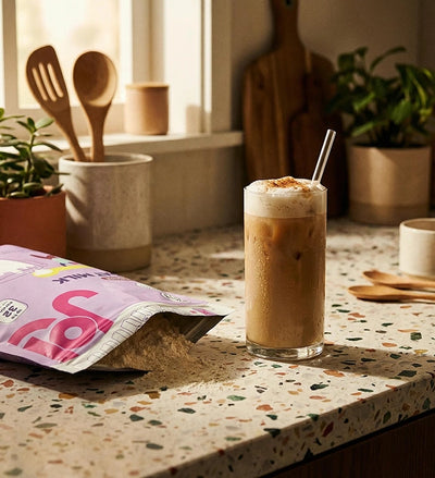 Glass of iced coffee made with oat milk powder, served on a kitchen countertop beside an open bag of plant-based milk mix.