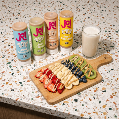 Four JOI plant bases in light blue, green, beige, and yellow bottles next to a glass of milk and fruit board.